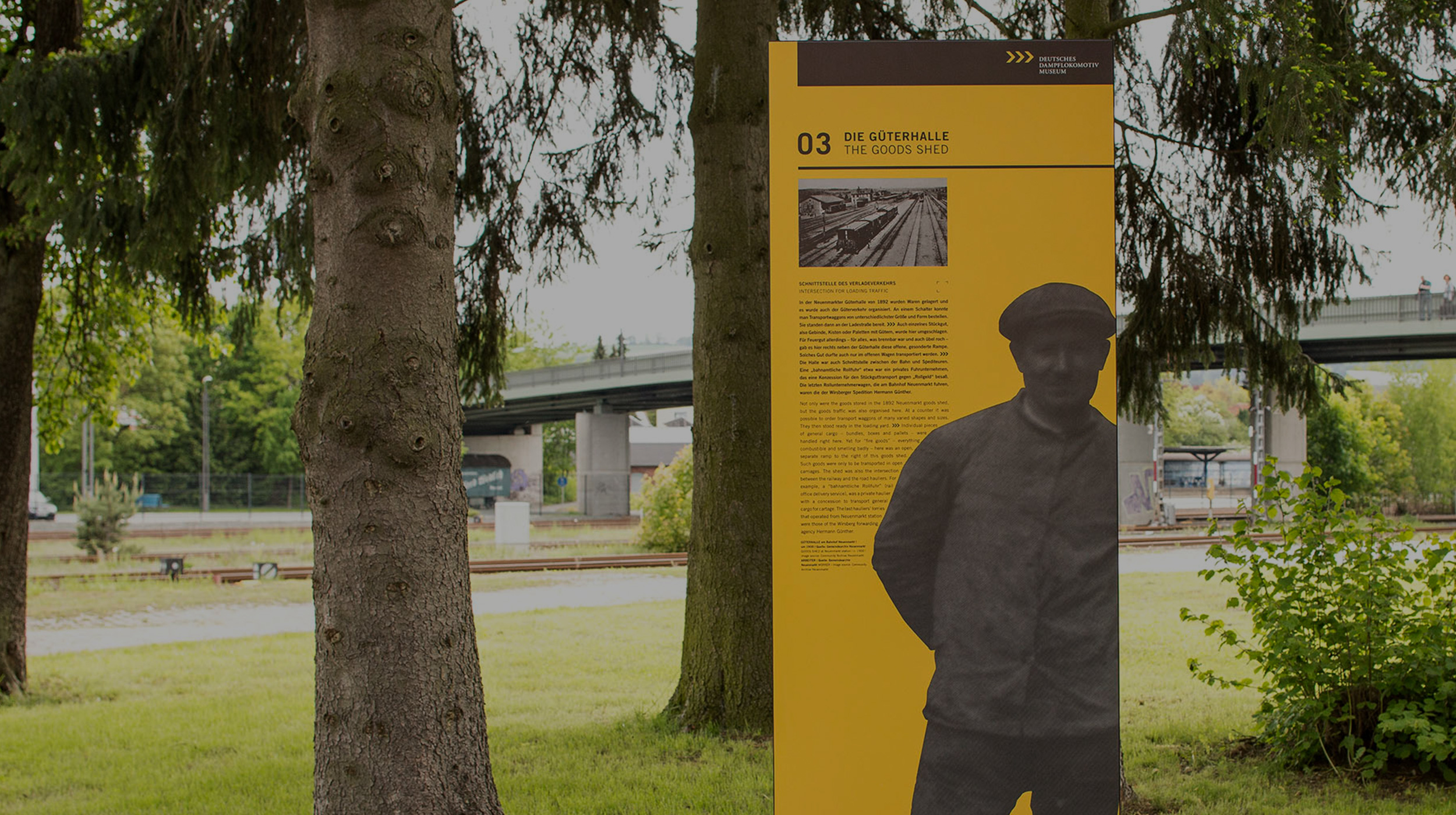A yellow information panel standing under a conifer in the railway town