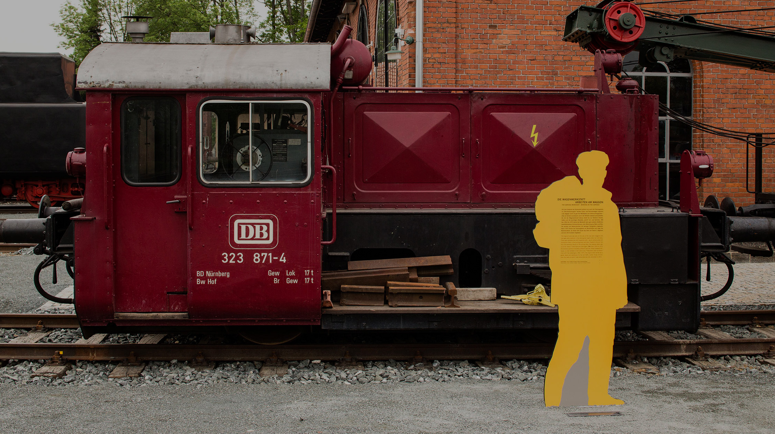 Eine lebensgroße CutOut-Figur mit aufgedrucken Informationen steht vor einer roten kleinen Diesellok